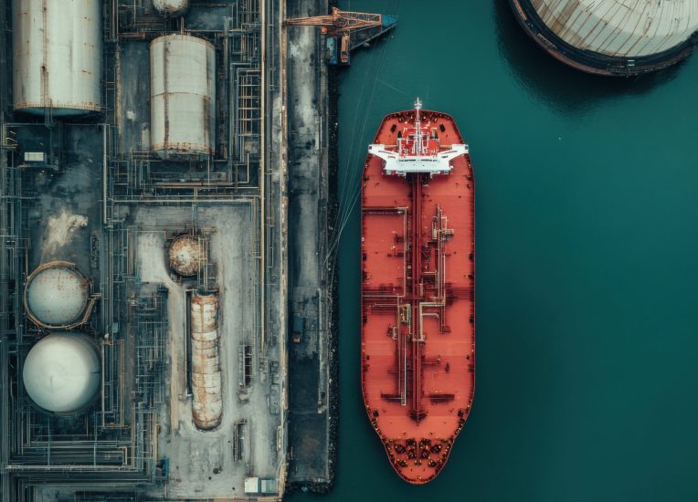 A large red cargo ship is docked at an industrial port surrounded by storage tanks and cranes. The scene captures the busy nature of maritime transport and trade activities.
